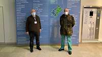 A soldier and another man standing in front of a floor plan of the hospital wearing masks and keeping a distance.