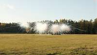 A dense smoke screen building up on a green field in front of woods.