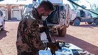 Ein Soldat steht an einem Tisch und reinigt seine Waffe, im Hintergrund ein Einsatzfahrzeug 