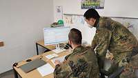 Ein Soldat erklärt einem anderen, der am Computer sitzt, das Arbeitsprogramm.