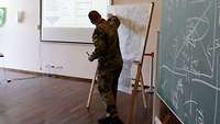 Ein Soldat steht neben einer aufgebauten Karte und erläutert, rechts hängt eine grüne Tafel mit Kreidezeichnungen.