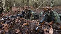Zwei mit Blättern getarnte Soldaten liegen mit ihren Waffen im Anschlag in einer Stellung im Wald.