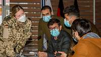 Vier Zivilisten diskutieren mit einer Soldatin, alle tragen einen Mund-Nasen-Schutz