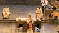 Ein Bischof mit Mitra steht vor einem steinernen Altar, dahinter sitzen weitere Geistliche.