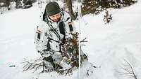 Ein Soldat in Schneetarnuniform hockt im Schnee. Vor ihm steckt ein weißer Skistock und ein Tannenzweig im Schnee.