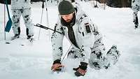 Ein Soldat in weißer Schneetarnuniform fährt mit dem Suchgerät über den weißen Schnee. 