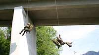 Zwei Soldaten hängen an Seilen, die an einer Betonbrücke über ihnen befestigt sind. 