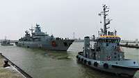 Ein graues Kriegsschiff vor einer Hafenpier, vorne und hinten je ein grauer Schlepper.