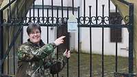 Weibliche Person zeigt auf ein Schild mit der Aufschrift "Fledermäuse willkommen" an einem Eisentor vor einem Gebäude