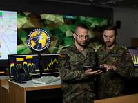 Zwei Soldaten stehen nebeneinander und schauen auf ein Tablet, hinter ihnen mehrere Computer und ein großes Wanddisplay