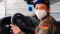 Eine Soldatin sitzt in Flecktarnuniform mit Mund-Nasenschutz-Maske am Lenkrad ihres Fahrzeugs 