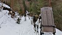 Soldaten in Schneetarnuniform laufen mit Skiern hintereinander einen verschneiten Waldhang hoch.