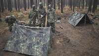 Soldaten stehen im Wald in der Dämmerung, vor und neben ihnen sind zwei Zwei-Mann-Zelte aufgebaut.