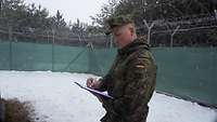 Ein Soldat steht mit einem Klemmbrett auf einer schneebedeckten Wiese und notiert sich etwas, im Hintergrund ein Zaun 
