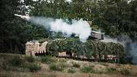 Ein Panzer steht vor einem Wald und verschießt eine kleine Rakete
