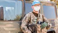 Ein Soldat mit Mund-Nasen-Schutz, blauem Basecap, Handschuhen, Weste und Gewehr steht an einem Fahrzeug