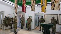 Fünf unterschiedliche Truppenfahnen hängen quer von einem grünen Holzgeländer herab, darunter stehen mehrere Soldaten.