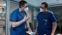 Zwei Männer in blauer Krankenhauskleidung sprechen miteinander. Vor ihnen liegt Sanitätsmaterial auf einem Tisch.