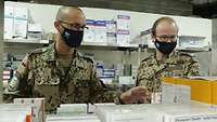 Zwei Soldaten stehen vor einem Regal mit Arzneimitteln. Beide tragen Mund-Nasen-Schutz.