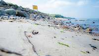 Ein Küstenstreifen mit Sand, Steinen und einem Hinweisschild „Militärischer Sicherheitsbereich“ auf einem Hügel.