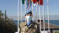 Eine Soldatin mit Mund-Nasen-Schutz steht vor acht verschiedenen Flaggen und dem Meer