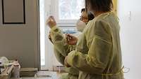 Zwei Frauen stehen im Impfraum in Schutzkleidung und halten je eine Spritze in der Hand.
