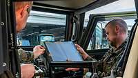 Zwei Soldaten der Bundeswehr montieren den Bildschirm eines Rechners in ein gepanzertes Militärfahrzeug ein.