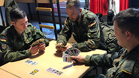 Drei Soldaten sitzen in der Stube an einem Schreibtisch und spielen Karten.
