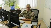 Eine Soldatin sitzt an einem Schreibtisch mit zwei Monitoren. Hinter ihr scheint Licht durch das Fenster