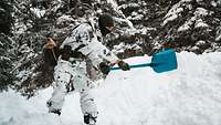 Ein bewaffneter Soldat in Wintertarnuniform schaufelt Schnee beiseite. Um ihn herum sind verschneite Nadelbäume.