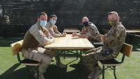 Vier Soldaten verschiedener Nationen mit Mund-Nasen-Schutz sitzen an einem Tisch