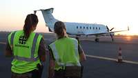 Zwei Soldatinnen des Sanitätspersonals stehen vor einem Rettungsflugzeug. Beide tragen gelbe Warnwesten