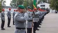 Mehrere Soldaten stehen auf einem Appellplatz in einer Reihe nebeneinander und grüßen militärisch.