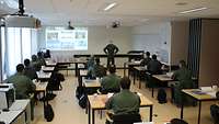 Die Teilnehmer des Waffenlehrerkurses sitzen in einem Lehrsaal und werden von einem Soldaten begrüßt.
