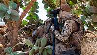 Ein Soldat sitzt im Busch und schaut durch einen Entfernungsmesser. Ein zweiter Soldat sitzt vor ihm mit seiner Waffe G22A2. 