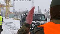 Im Vordergrund steht ein Soldat, der ein Fahrzeug einweist, im Hintergrund Häuser im Schnee