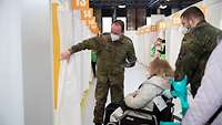 Ein Soldat hält den Vorhang einer Impfkabine, damit ein weiterer Soldat eine Frau in einem Rollstuhl hineinschieben kann