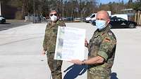 Zwei Soldaten stehen am neuen Standort und halten eine Urkunde.