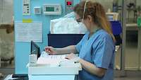 Eine Ärztin in Krankenhauskleidung steht auf der Intensivstation an einer Theke und schreibt in eine Tabelle