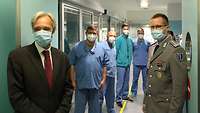 Ein Mann im Anzug, ein Soldat im Dienstanzug und vier Männer in Krankenhauskleidung blicken in eine Kamera