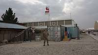 Ein Soldat steht vor mehreren Containern. Hinter ihm wehen auf einem der Container zwei rot-weiße Flaggen