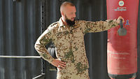Ein Soldat hält an seinem linken ausgestreckten Arm ein Gewicht und macht Übungen. Im Hintergrund hängt ein roter Boxsack