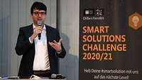 Ein Mann spricht in ein Mikrofon und gestikuliert, daneben ein Banner mit der Aufschrift "Smart Solutions Challenge 2020/21"
