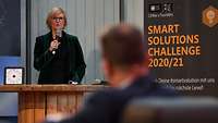 Eine Frau spricht in ein Mikrofon, daneben ein Banner mit der Aufschrift "Smart Solutions Challenge 2020/21"