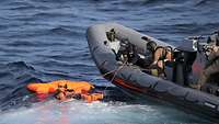 Ein graues Schlauchboot in See; daneben eine angeleihnte Person im Wasser, die eine lebensgroße orangene Puppe hält.