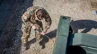 Ein Soldat steht gebeugt mit einer Metallstange in den Händen vor einem Gabelstapler und kratzt Sand ab