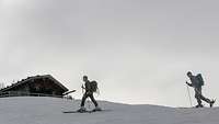 In einer Berglandschaft marschieren zwei Soldaten auf Tourenskiern an einer Almhütte vorbei.