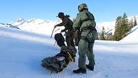 Zwei Soldaten in Pilotenanzug stehen im Schnee neben einer Trage, auf der ein Verletzter liegt.