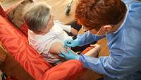 Eine grauhaarige Dame wird von einer Frau mit blauem Kittel in den Arm geimpft.