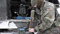 Ein Soldat kniet vor der Heckklappe eines Panzers. Er legt in diesen einen Munitionsgurt für das Maschinengewehr